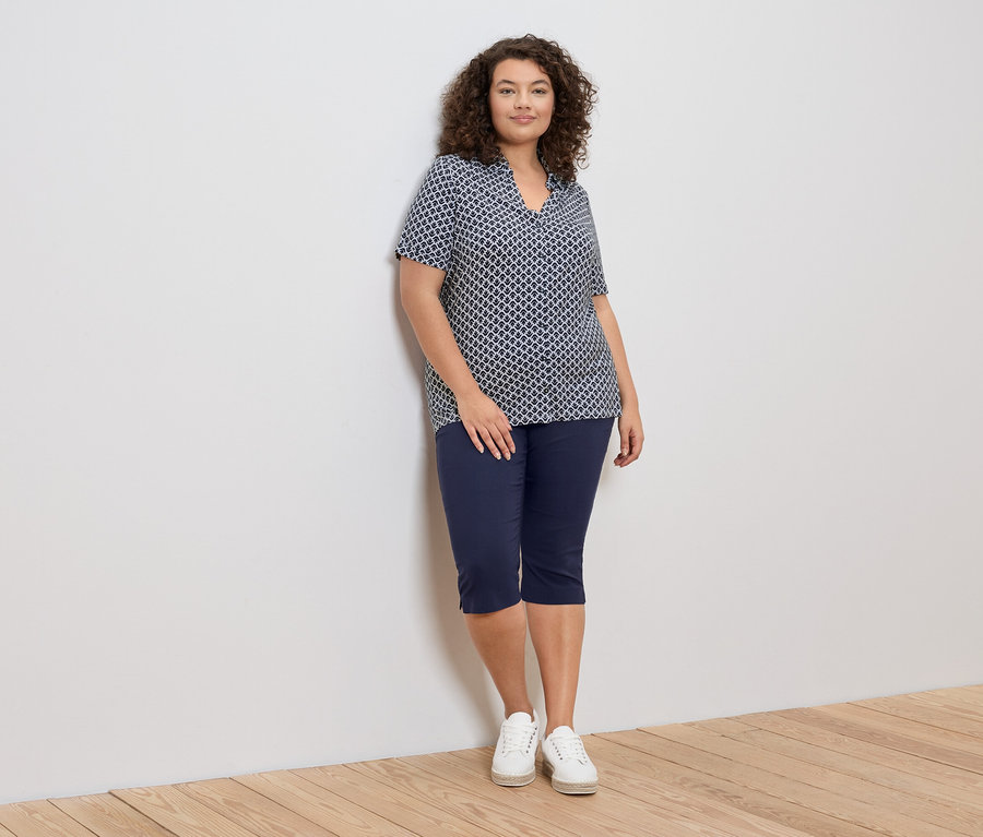 Egy göndör hajú nő a falnak támaszkodik, kék-fehér mintás blúzt, kék capri nadrágot és fehér tornacipőt visel.
