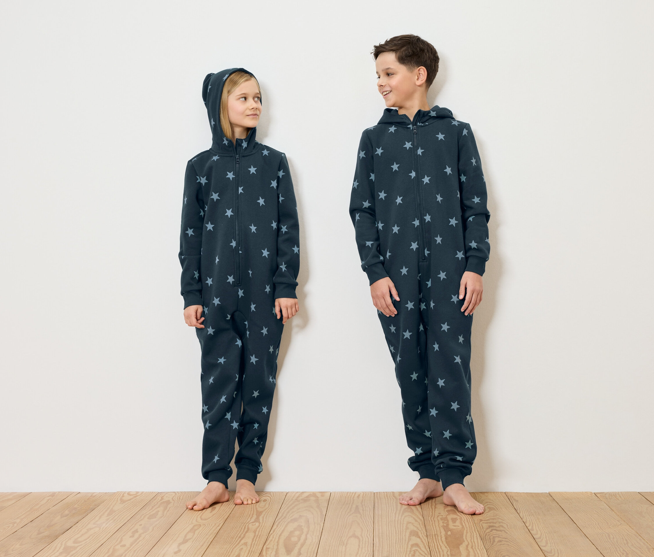 Egy lány és egy fiú mezítláb állnak egy fehér fal előtt, csillagmintás, sötétkék pizsamában.