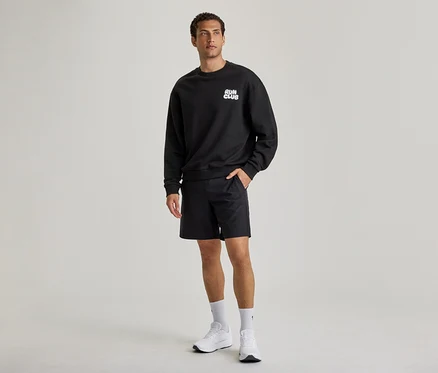 Egy férfi fekete szabadidőfelsőt visel „RUN CLUB” felirattal, fekete rövidnadrágot és fehér sportcipőt. Bal keze a rövidnadrág zsebében van.