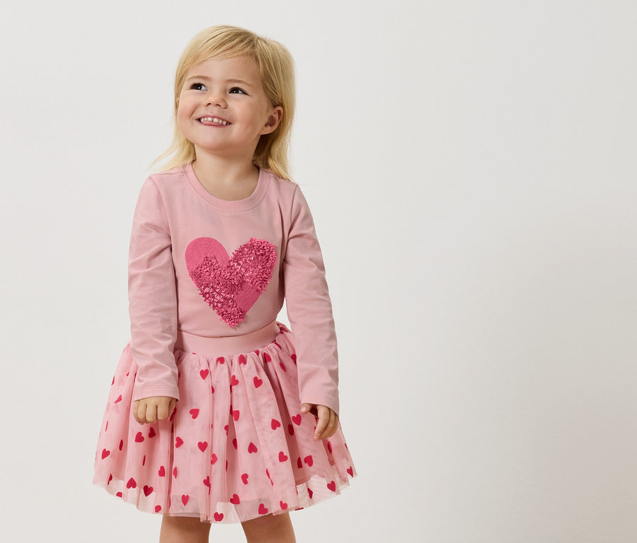 Egy kis szőke hajú lány rózsaszín, hosszú ujjú felsőt visel, nagy, rózsaszín szívvel, és rózsaszín tüllszoknyát piros szívekkel.