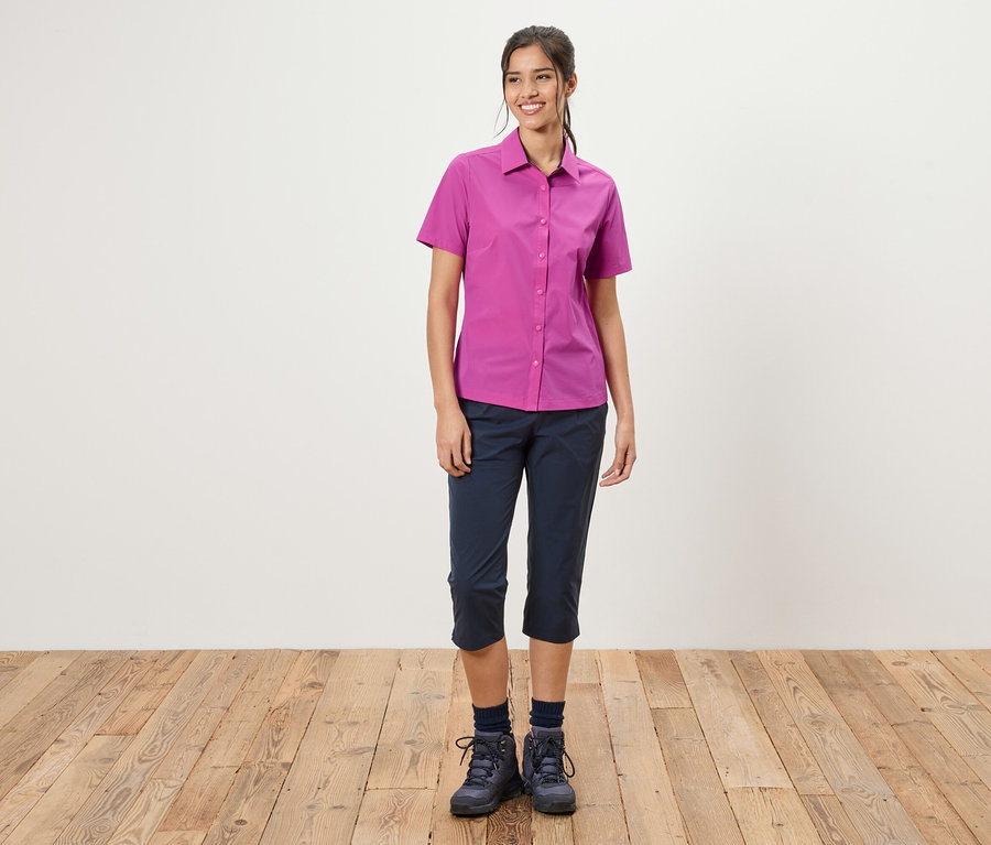 Egy nő áll egy fehér fal előtt. Rózsaszín inget, kék háromnegyedes nadrágot és túrabakancsot visel.