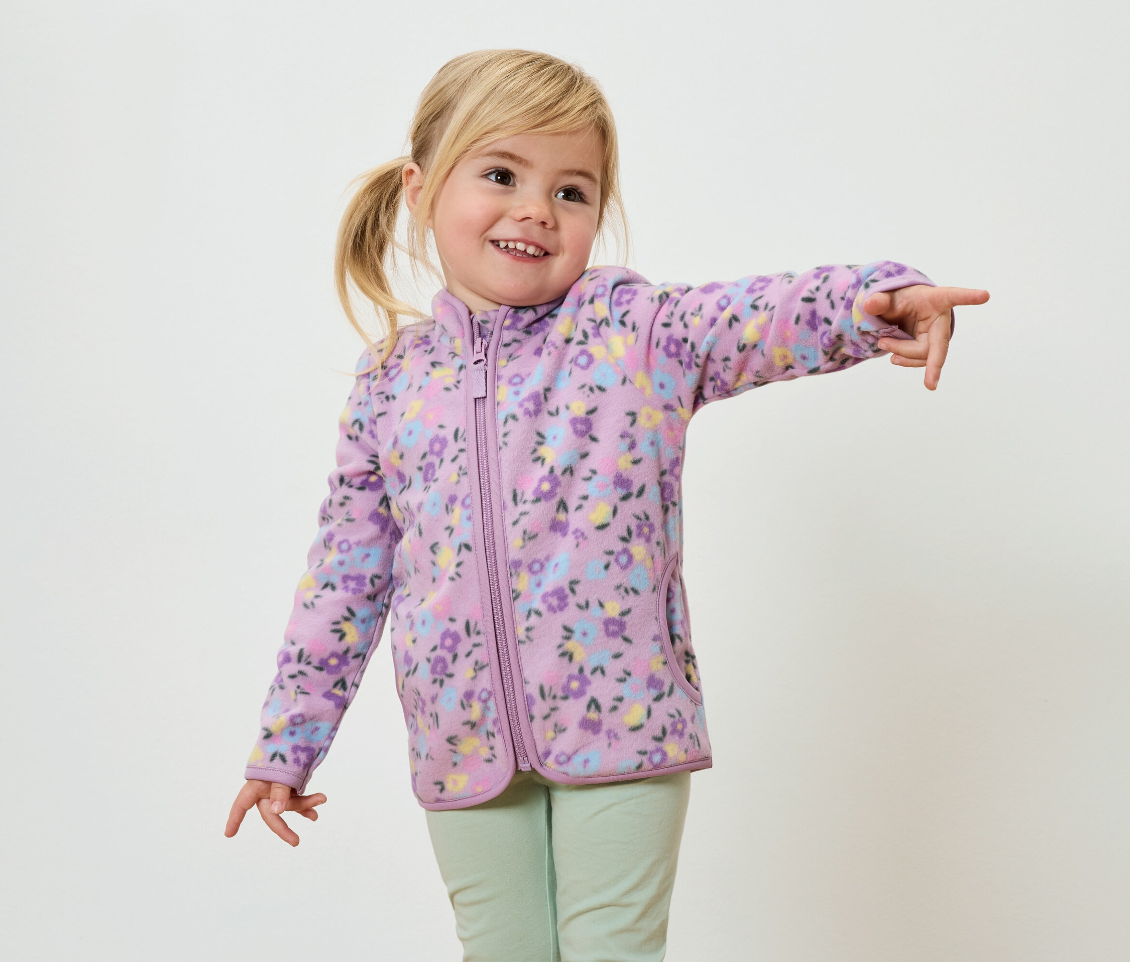 Egy mosolygó, szőke copfos kislány lila, virágmintás fleece dzsekiben és világoszöld nadrágban mutat az ujjával.