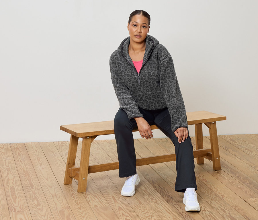 Nő ül egy fa padon, leopárdmintás szürke fleece pulóvert, rózsaszín felsőt, sötét nadrágot és fehér tornacipőt visel.