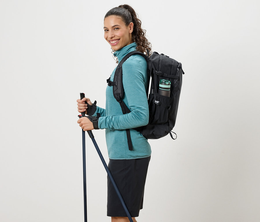 Türkiz garbót nyakú pulóvert viselő nő, fekete hátizsákkal és túrabotokkal.