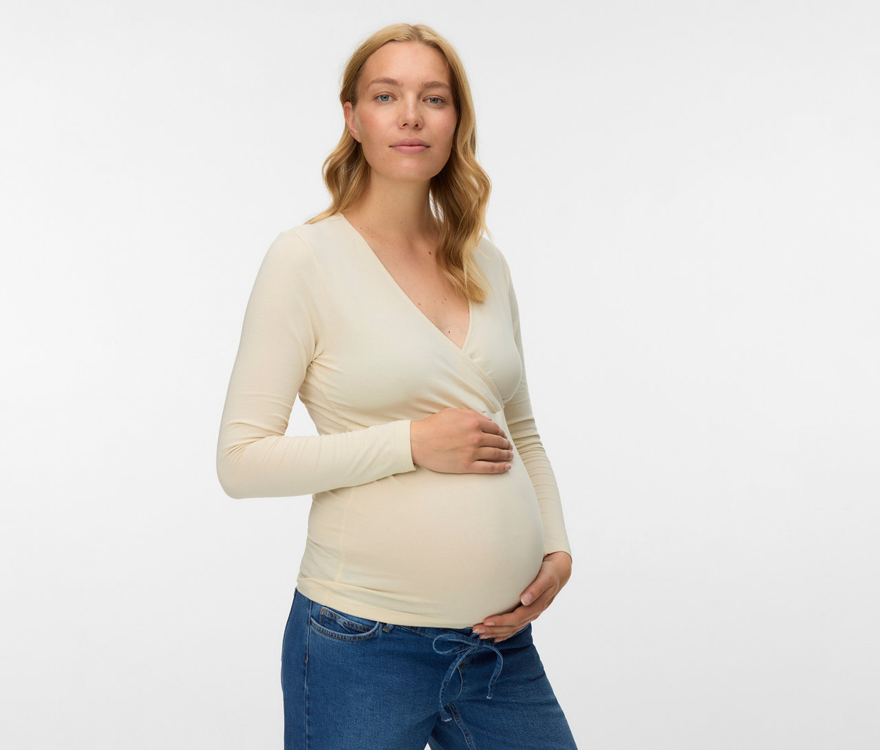 Terhes nő bézs színű, 2 db-os MAMALICIOUS kismama hosszú ujjú felsőben és farmerban, a hasát fogva.