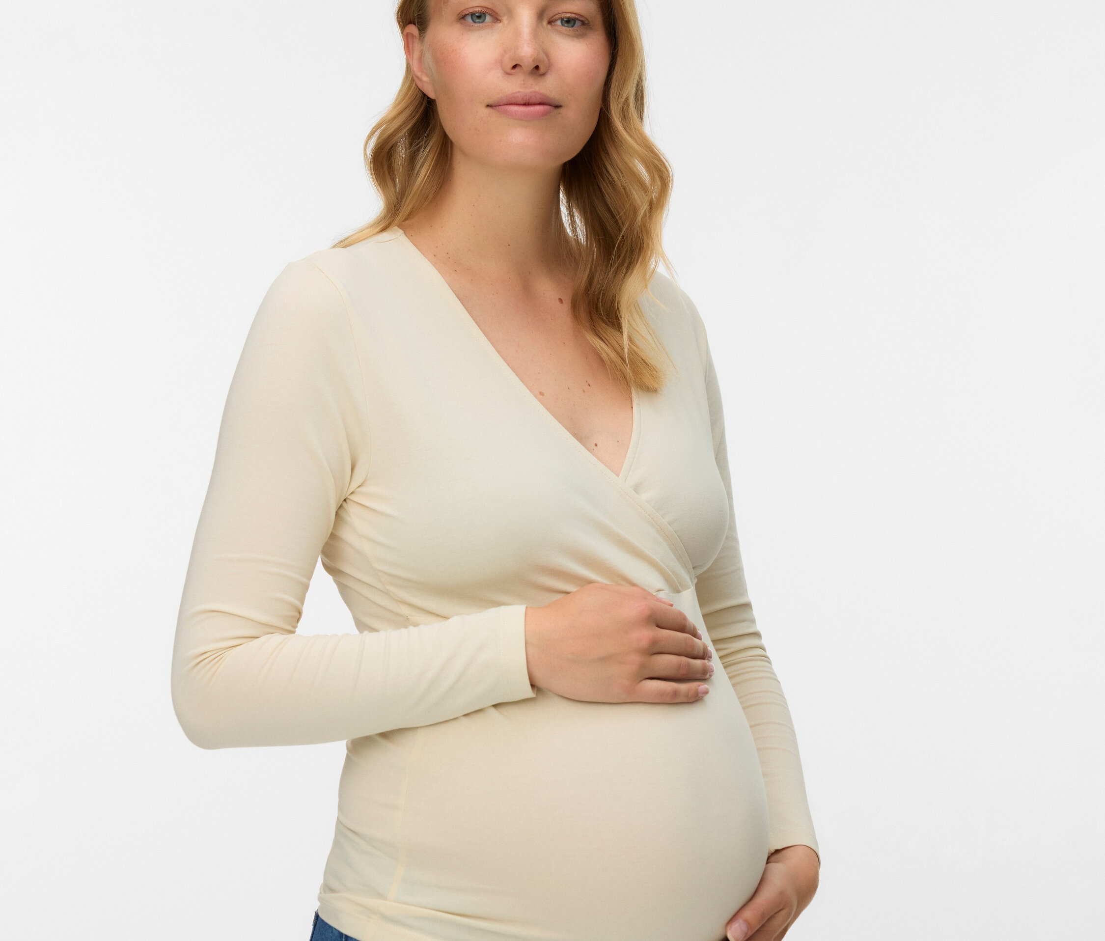 Egy várandós nő szőke, hullámos hajjal, hosszú ujjú átlapolt blúzt és farmert visel.