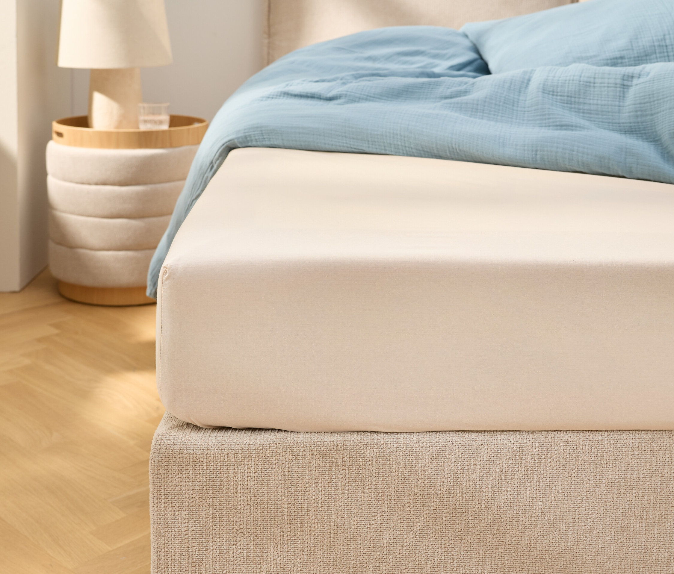 Közeli kép egy bézs színű gumis lepedőről egy világoskék ágyneművel ellátott ágyon. Mellette egy éjjeliszekrény áll egy lámpával.