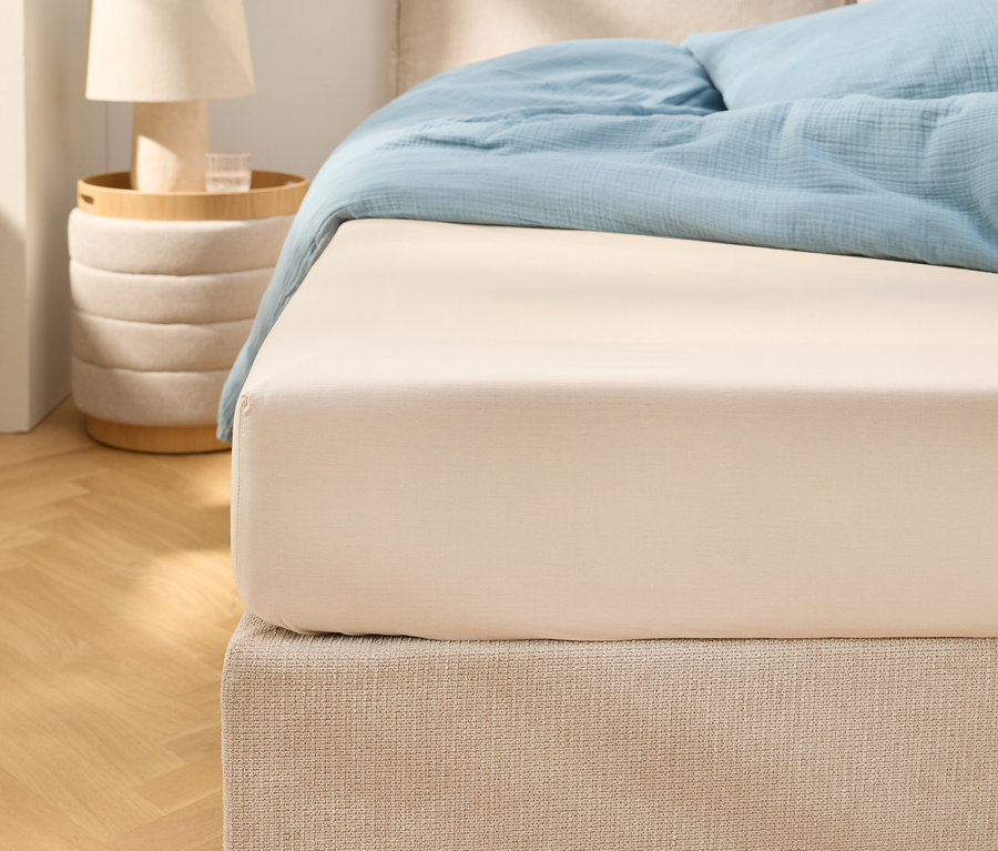 Közeli kép egy bézs színű gumis lepedőről egy világoskék ágyneművel ellátott ágyon. Mellette egy éjjeliszekrény áll egy lámpával.