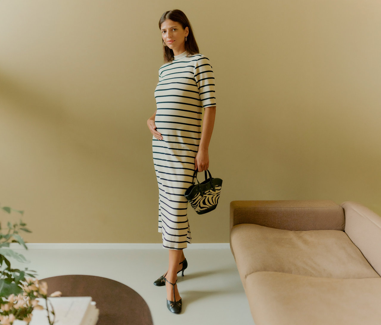 Egy nő csíkos MAMALICIOUS kismama midiruhában kézitáskát tart és a hasát fogja. Fekete cipőt visel.