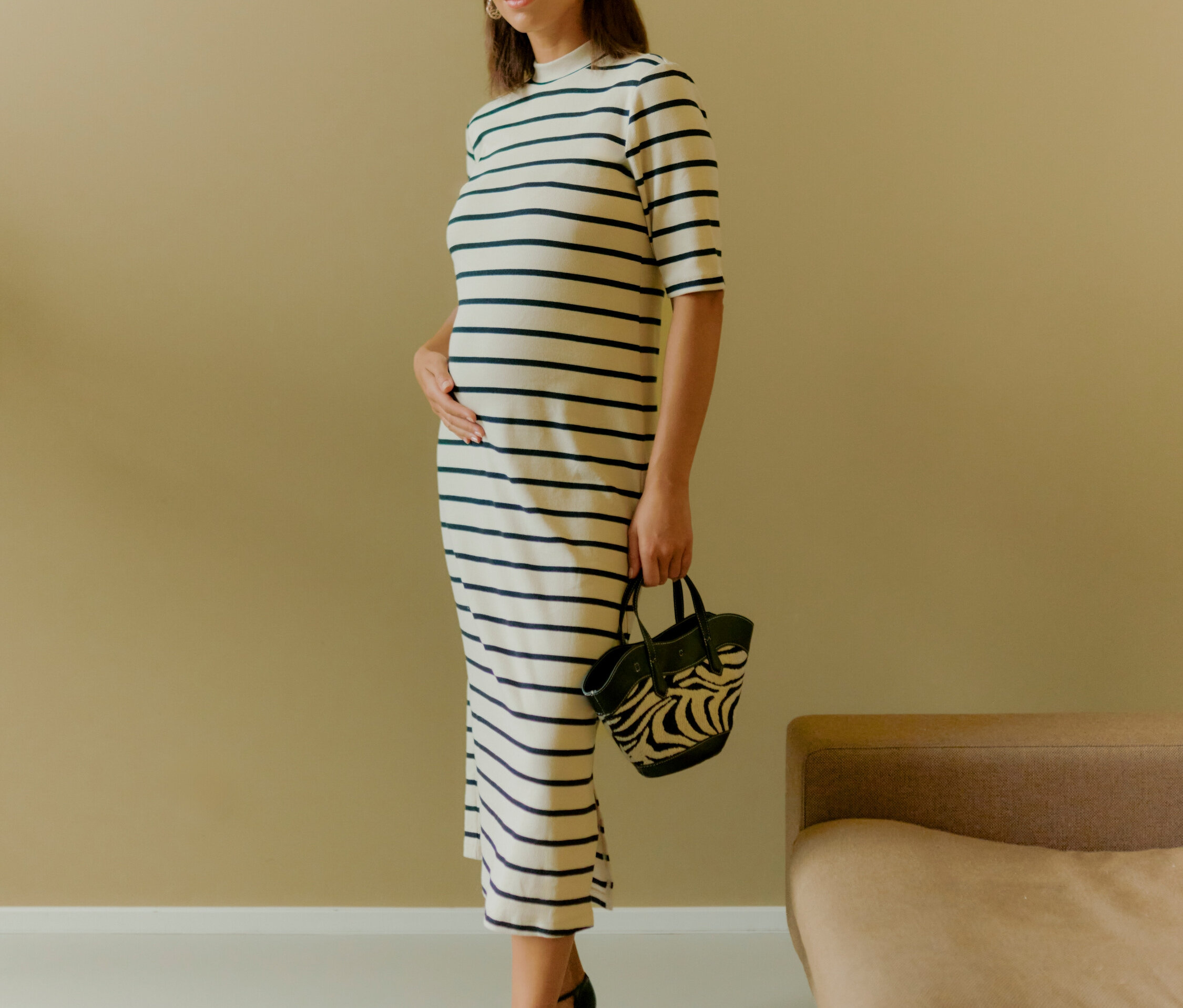 Egy nő áll és egy zebra mintás kézitáskát tart. Csíkos ruhát és fekete cipőt visel.