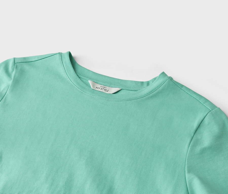 Egy világos türkiz színű póló közeli képe, rajta egy "TCHIBO MY STYLE" feliratú címkével.