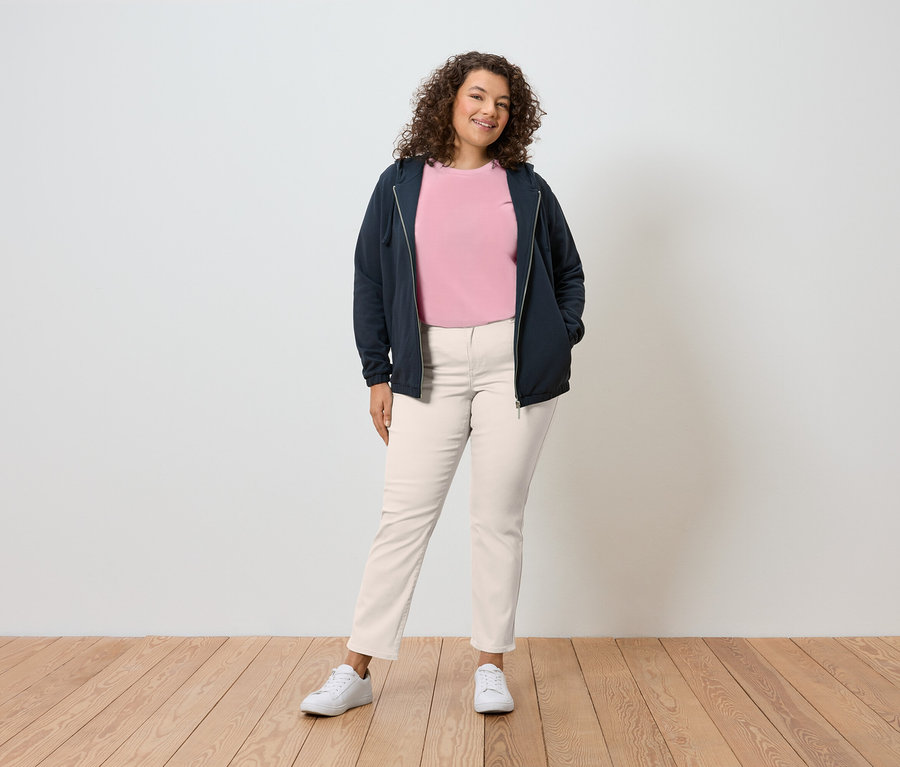 Egy göndör hajú nő áll a fal előtt, sötétkék kapucnis pulóvert, rózsaszín pólót, krémszínű nadrágot és fehér tornacipőt visel.