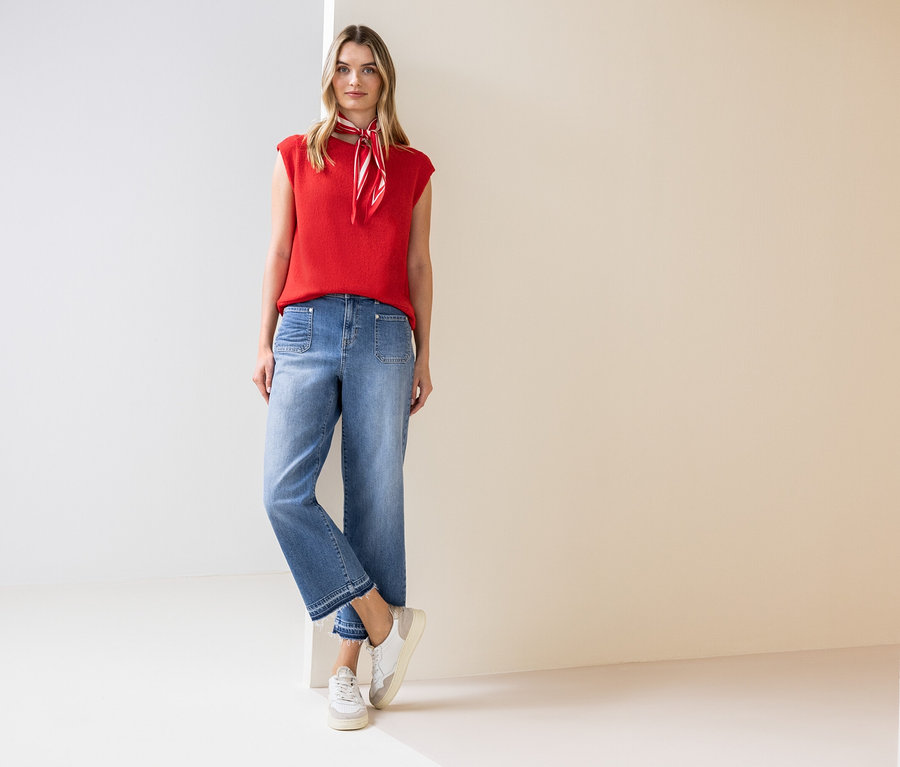 Egy nő áll, piros felsőt, nyakláncot, farmert és tornacipőt visel.