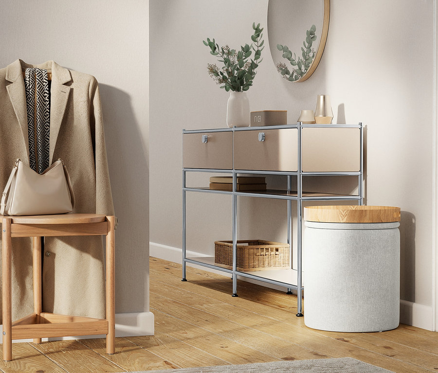 Egy bézs kabát lóg a falon egy mellette lévő zsámolyon, rajta egy kézitáskával. Mellette egy fiókos polc és egy kerek ülőke áll.