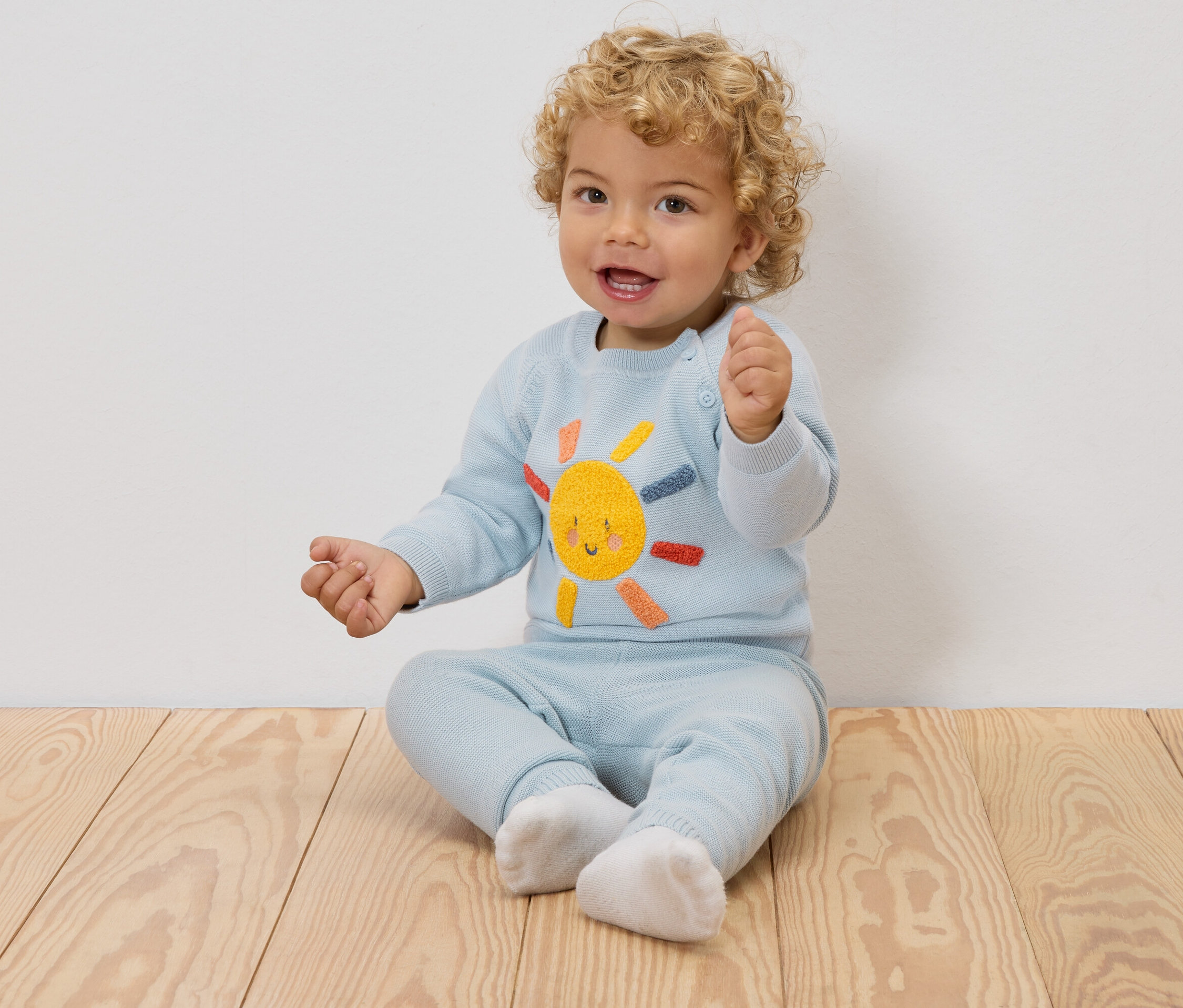 Egy mosolygó, szőke fürtös baba ül egy fapadlón, világoskék pulóvert visel, hímzett napocskával, és hozzáillő nadrágot.