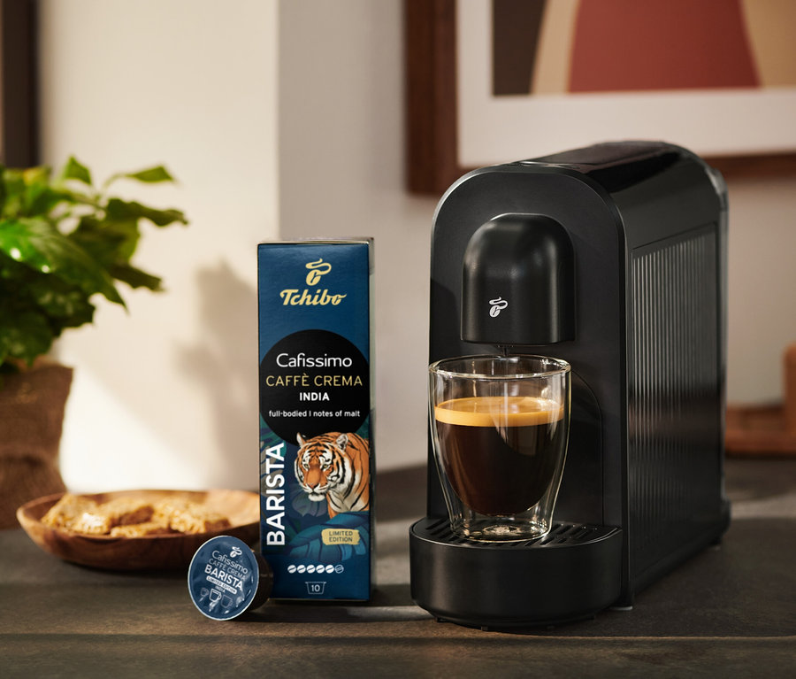 Fekete Tchibo Cafissimo kávéfőző egy pohár kávéval, mellette egy csomag Tchibo Cafissimo Caffè Crema India kávékapszula és egyetlen kapszula.