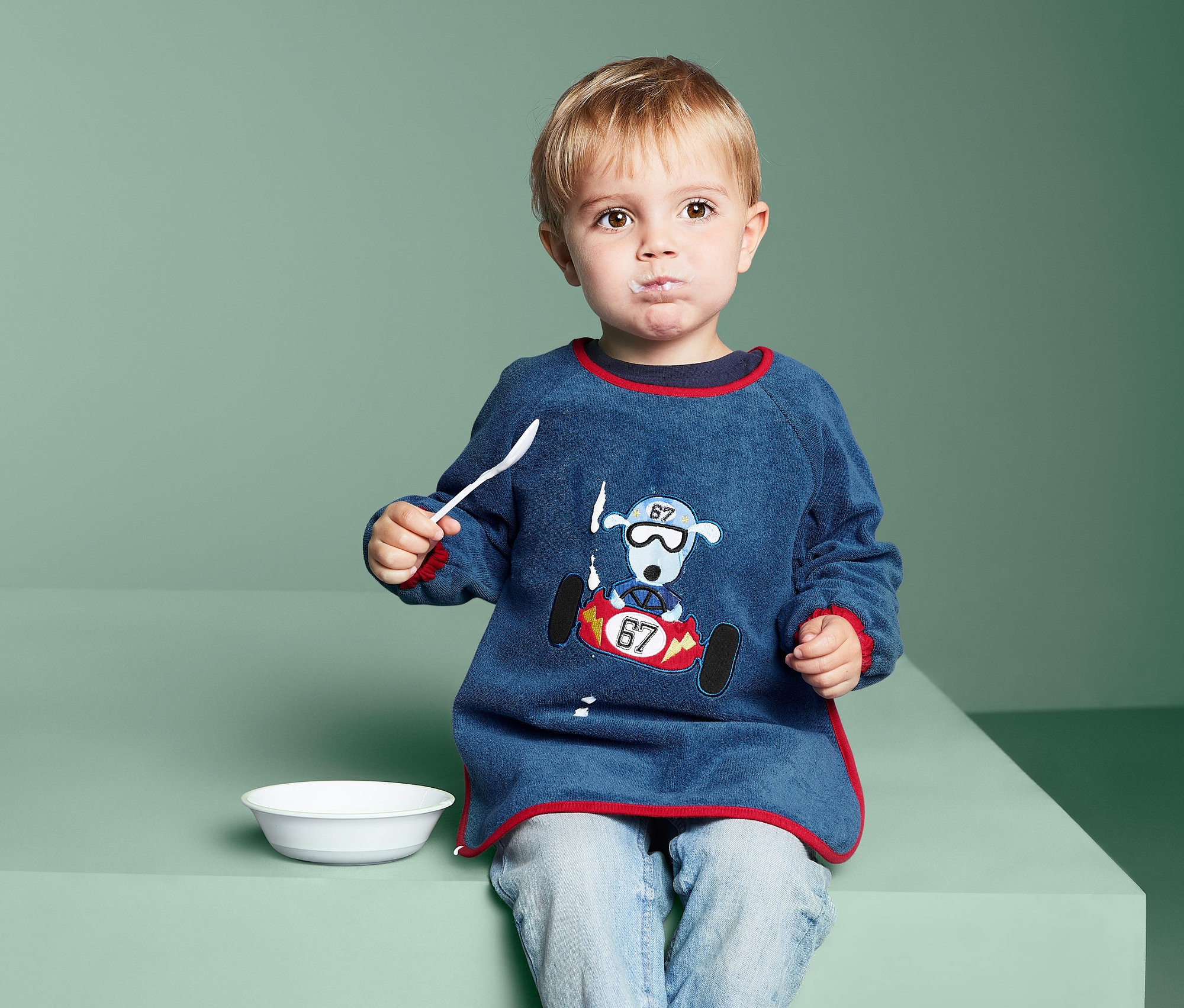 Egy gyermek kanállal eszik a 2 ujjas előke szettben lévő előkét viselve.