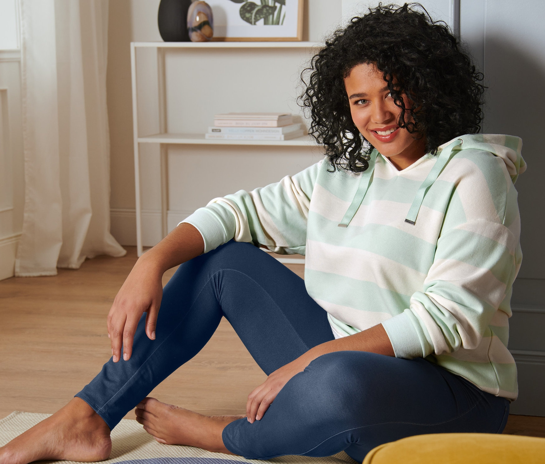 Egy nő ül a padlón, és egy menta színű női kapucnis szabadidőfelsőt és egy farmer hatású női leggingst visel.