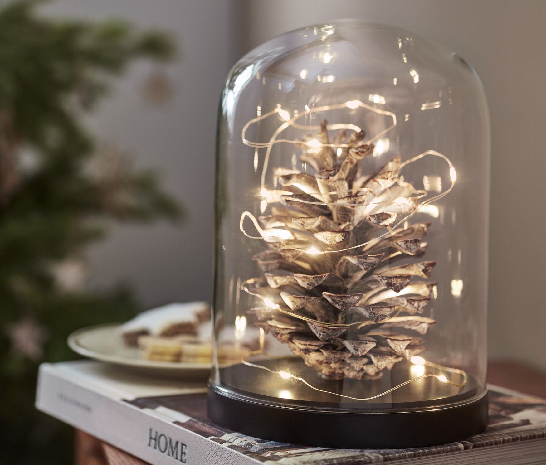 Egy üvegburában lévő toboz LED-es mikro égősorral, 20 cseppel áttekerve egy könyvön áll.