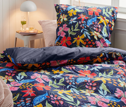 Virágmintás perkál ágyneművel ellátott ágy, beleértve a dupla, normál és extra méreteket. Az éjjeliszekrényen lámpa áll.