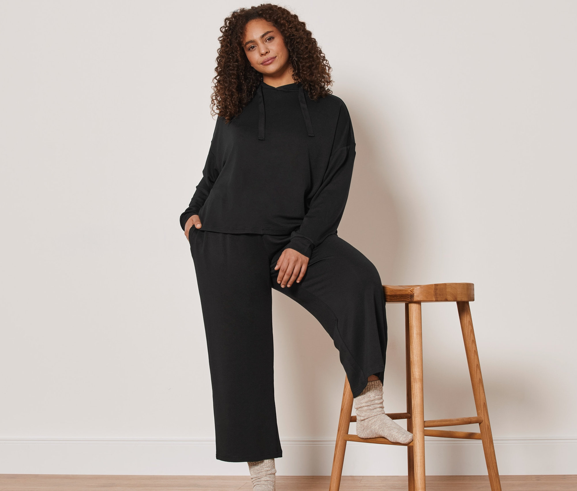 Nő pózol egy fa széknek dőlve, fekete 7/8-os szabadidő szettben.