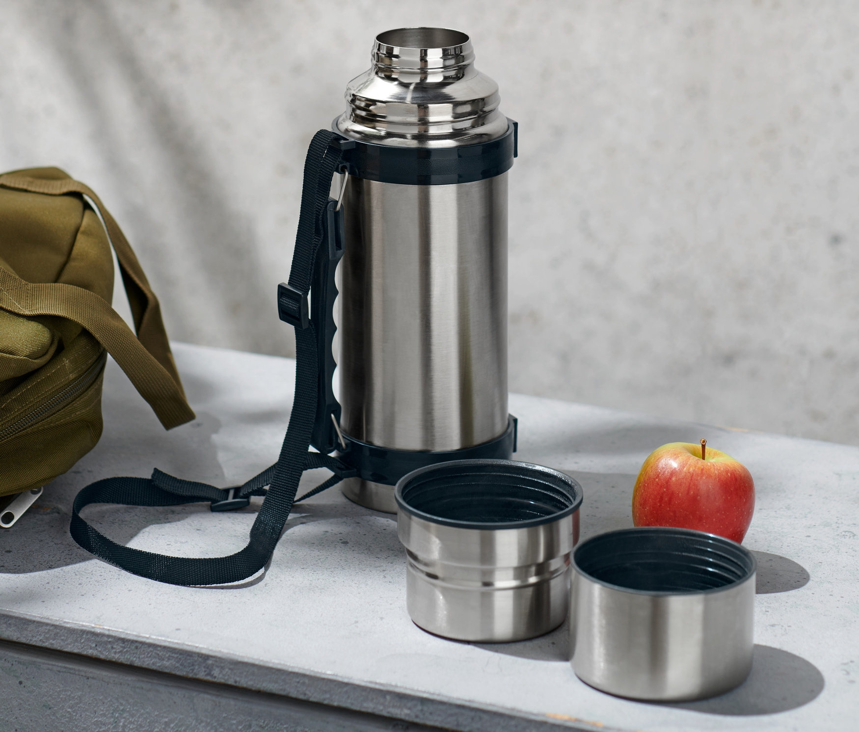 Egy ezüst termosz két pohárral, egy olívazöld táska és egy alma fekszik egy szürke felületen.