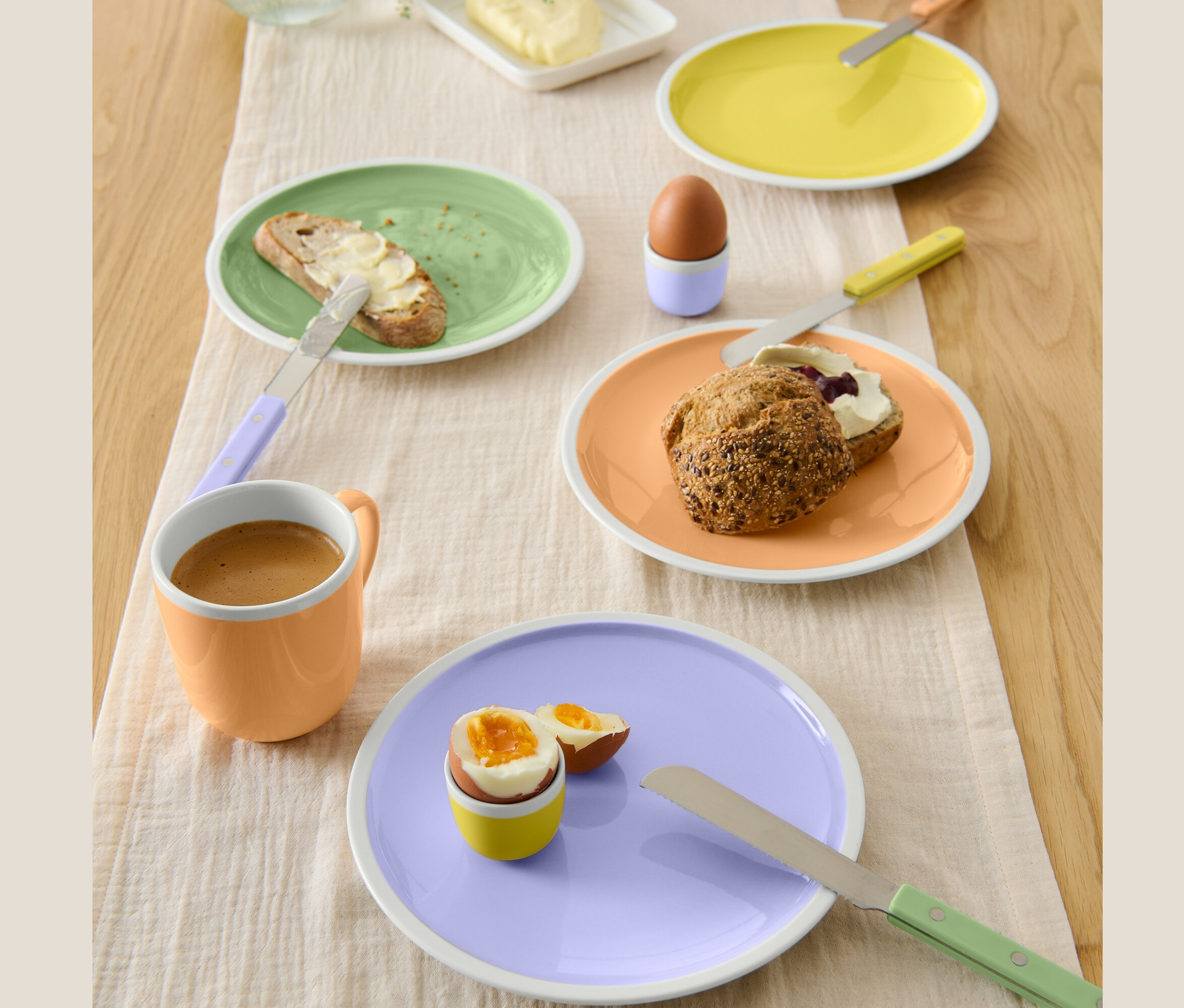Az asztalon négy különböző színű porcelán tányér található reggelivel. A tányérokon vajas kenyér, tojás, dzsemes és sajtos zsemle.