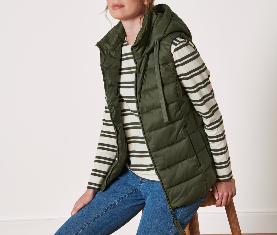 Nő ül egy széken, és kapucnis, olíva színű steppelt mellényt visel.