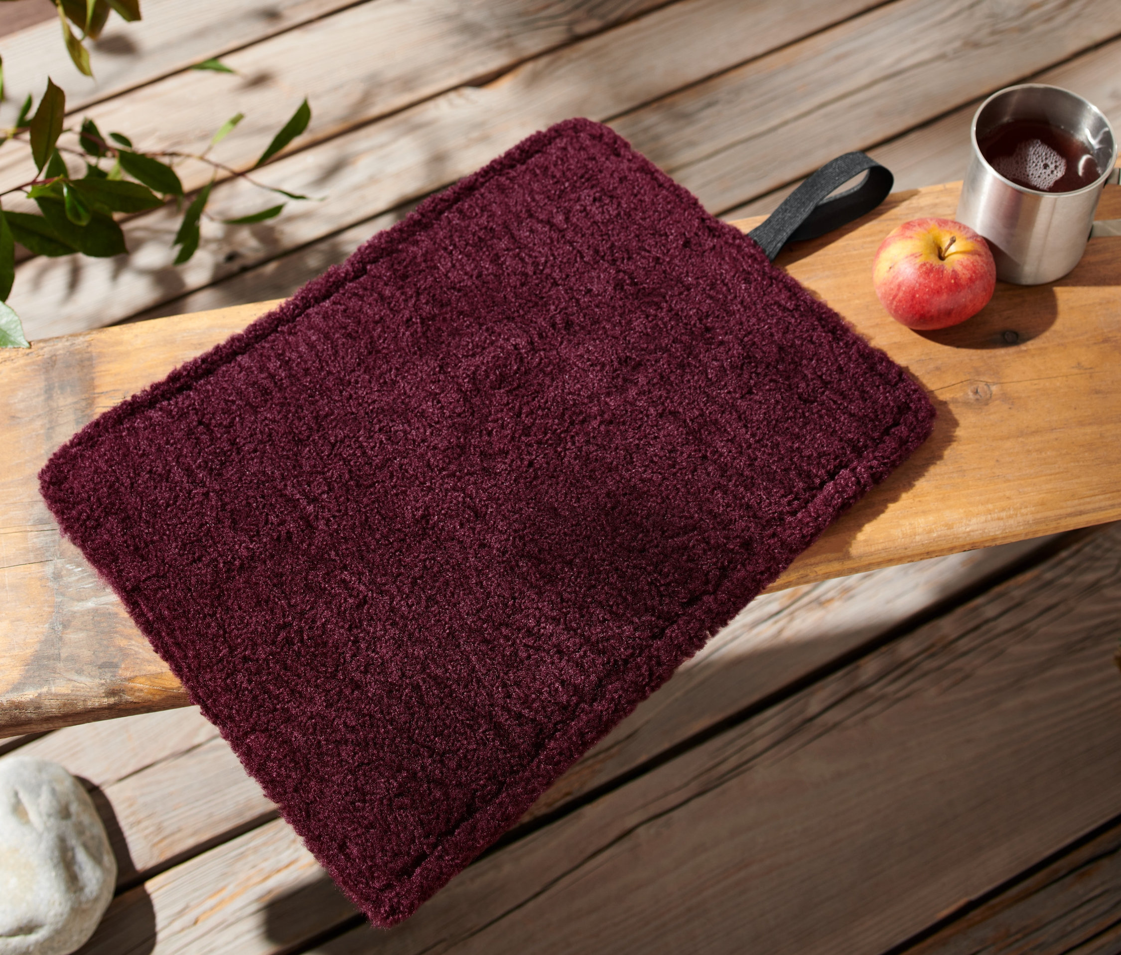 Bordó színű összehajtható termo ülőpárna egy fa padon fekszik egy alma és egy csésze mellett.