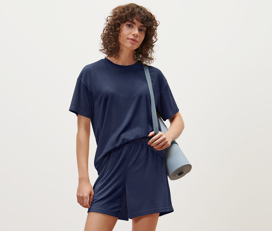 Nő visel sötétkék frottír felsőt és rövidnadrágot, a vállán pedig egy jógamatracot.