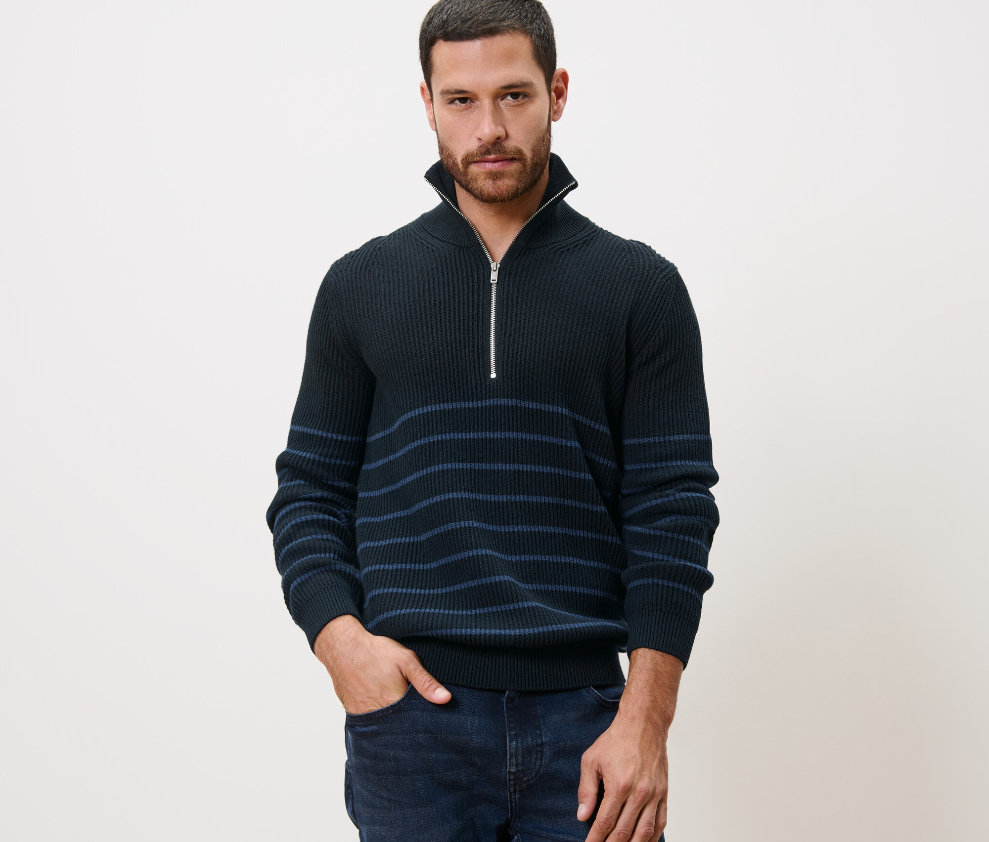 Férfi pózol csíkos, sötétkék állónyakú pulóverben, keze a farmerzsebében.