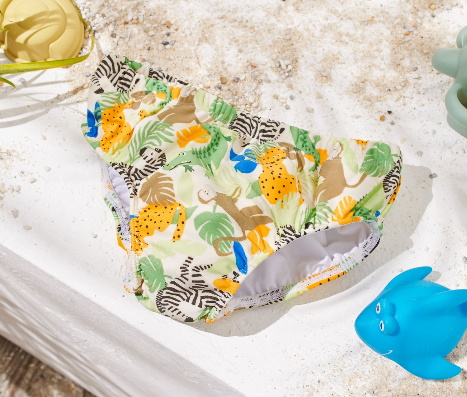 Állatmintás baba úszópelenka fekszik egy fehér felületen homokozó játékok mellett.