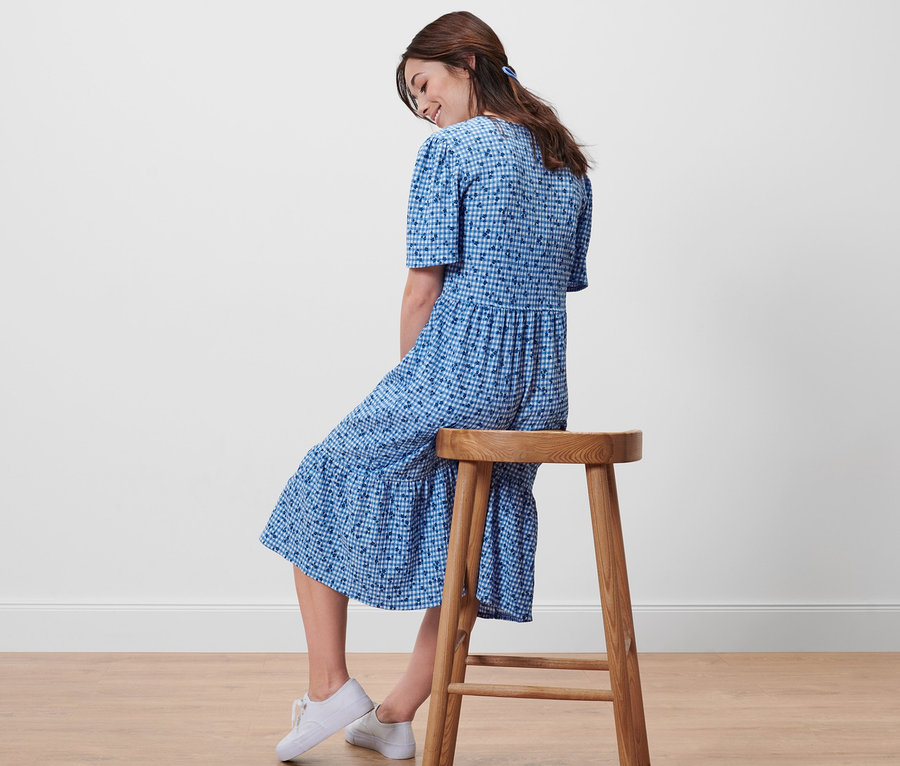 Nő ül oldalra egy fa székleten, kék, mintás ruhában.