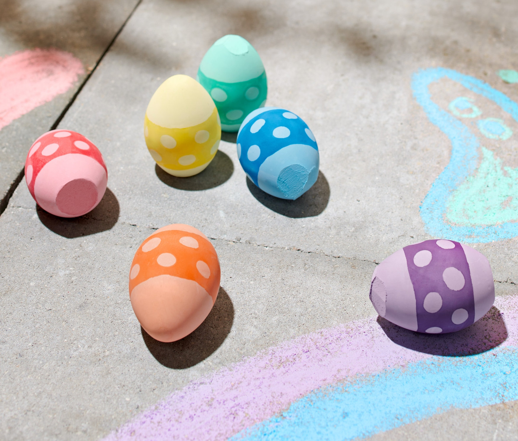 Hat színes, tojás alakú utcai kréta fekszik egy járdán krétarajzok mellett.