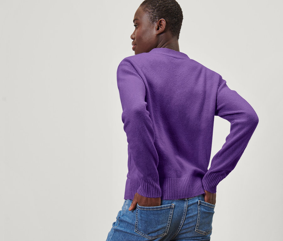 Egy nő háttal pózol, keze a farmerzsebében. Lila, gyapjú kötött kardigánt visel.