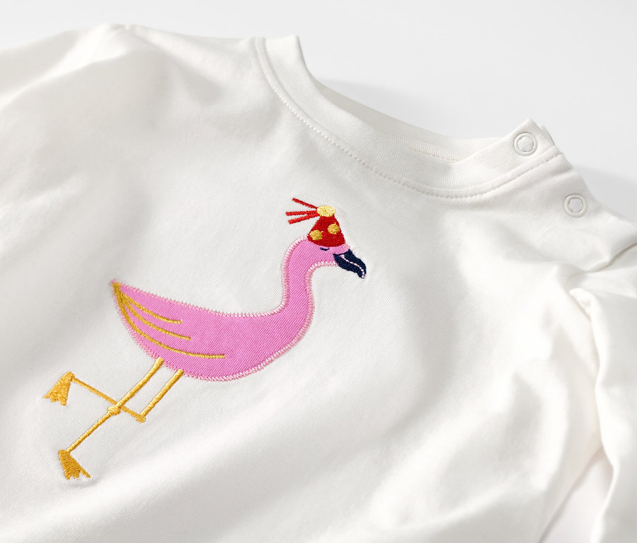 Egy fehér, hosszú ujjú gyerek póló részlete, hímzett flamingóval és születésnapi sapkával.