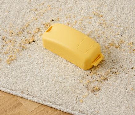 Egy sárga morzsaseprű fekszik egy bézs szőnyegen szétszórt morzsákkal.