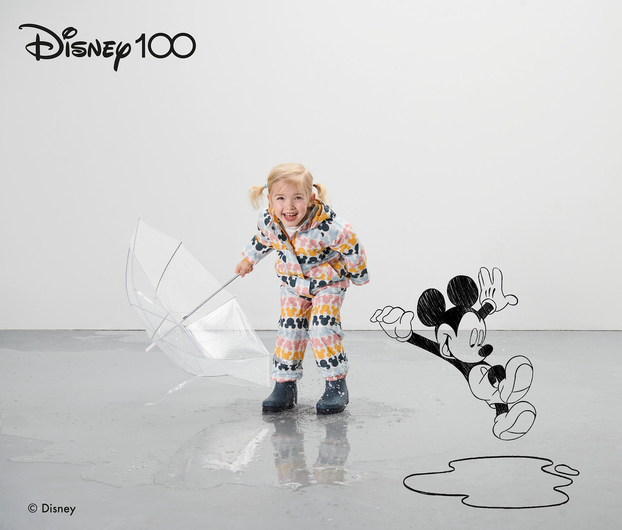 Egy kislány színes »Micky« termo esőkabátban áll egy tócsában, átlátszó esernyőt tartva.