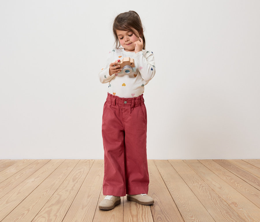 Kislány tart egy fa fényképezőgépet, és ránéz. Nadrágot és hosszú ujjú inget visel.