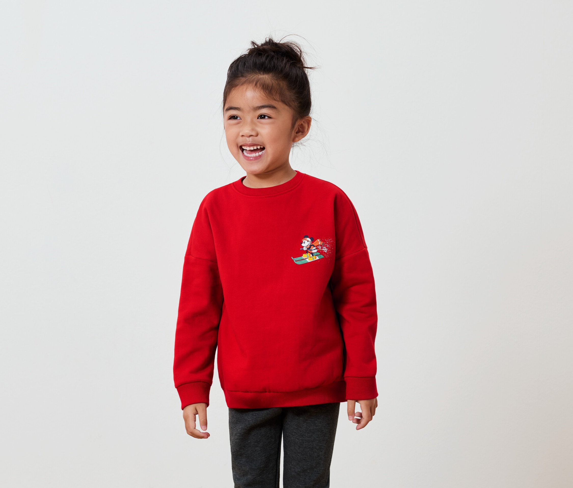 Egy mosolygó lány piros pulóvert visel, rajzfilmfigurával a mellkasán.