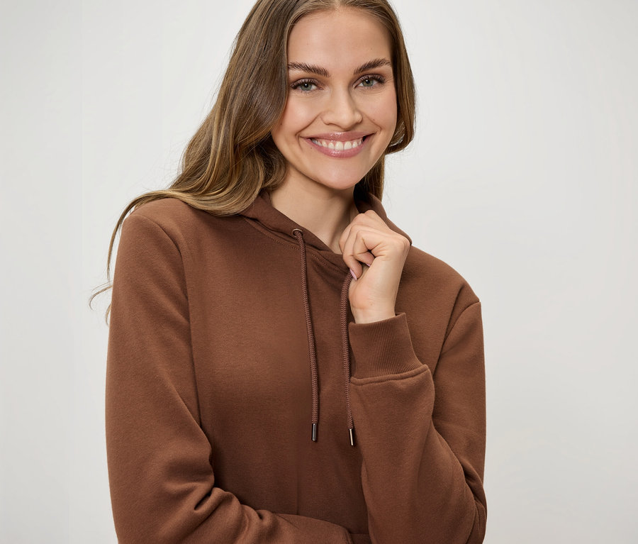 Mosolygó nő barna, húzózsinóros kapucnis pulóverben, egyik kezével a gallérját fogja.