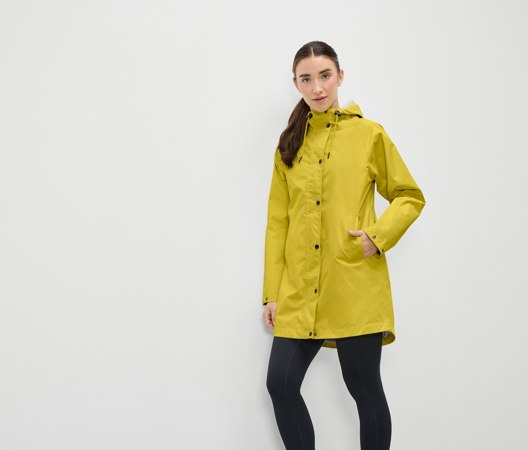 Egy nő sárga esőkabátban, kapucnival és fekete leggingsben áll egy fehér fal előtt.