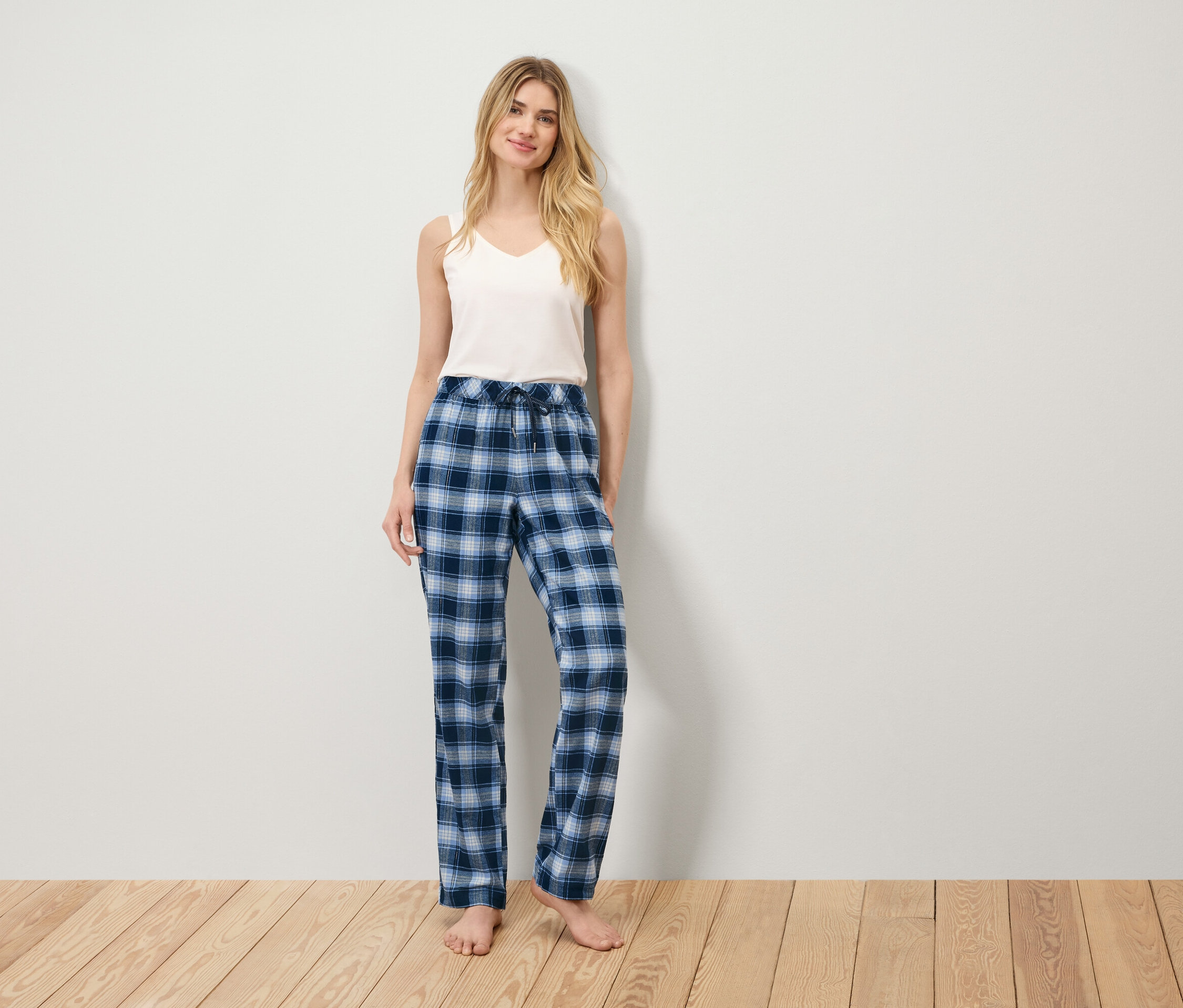 Egy nő áll egy fapadlón, fehér trikót és kék kockás pizsamanadrágot visel.