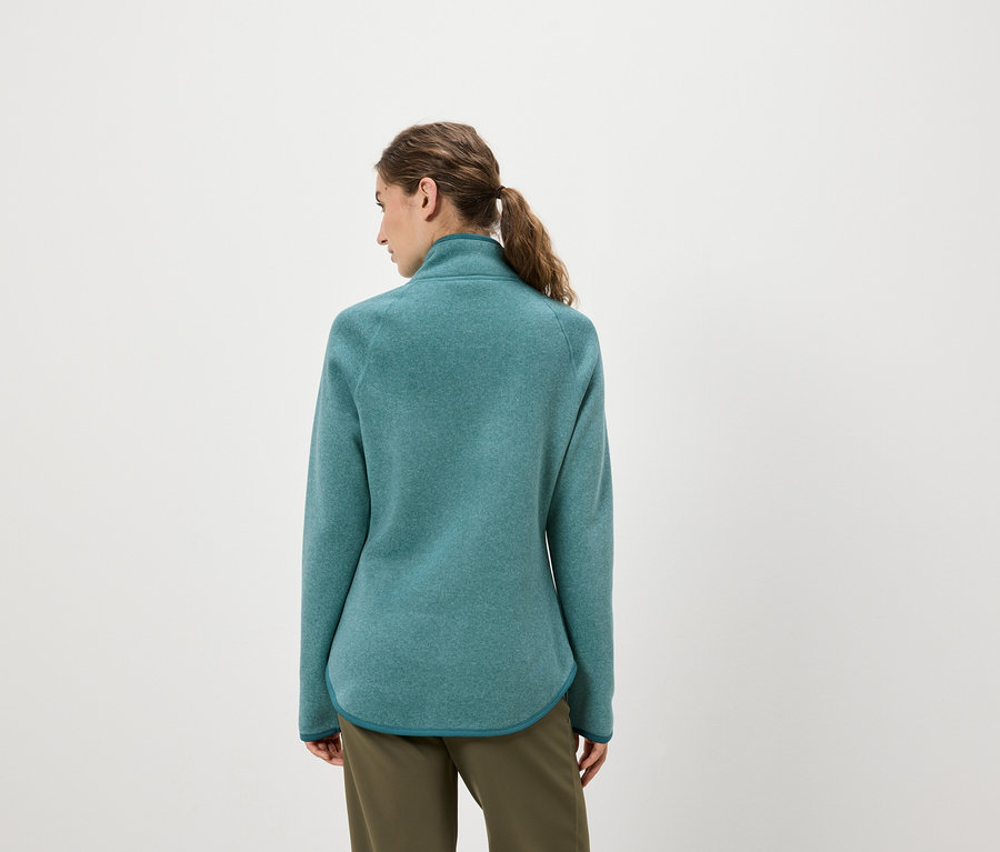 Egy nő hátulról nézve türkizkék polár pulóvert és olívazöld nadrágot visel.