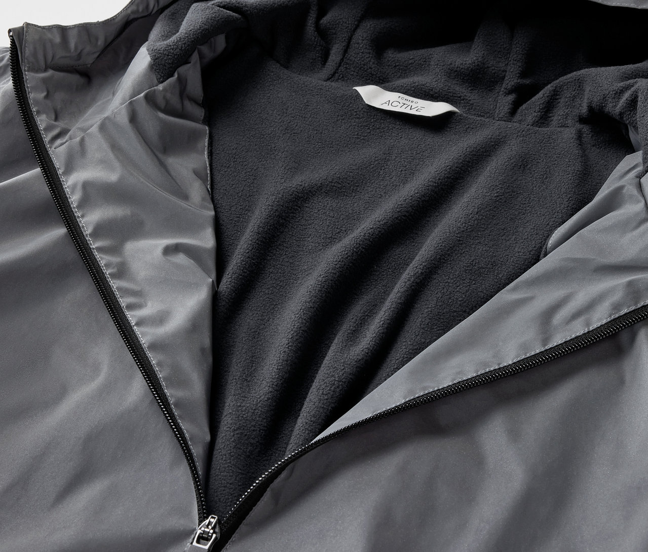 Egy szürke, fényvisszaverő sportkabát részlete, nyitott cipzárral.