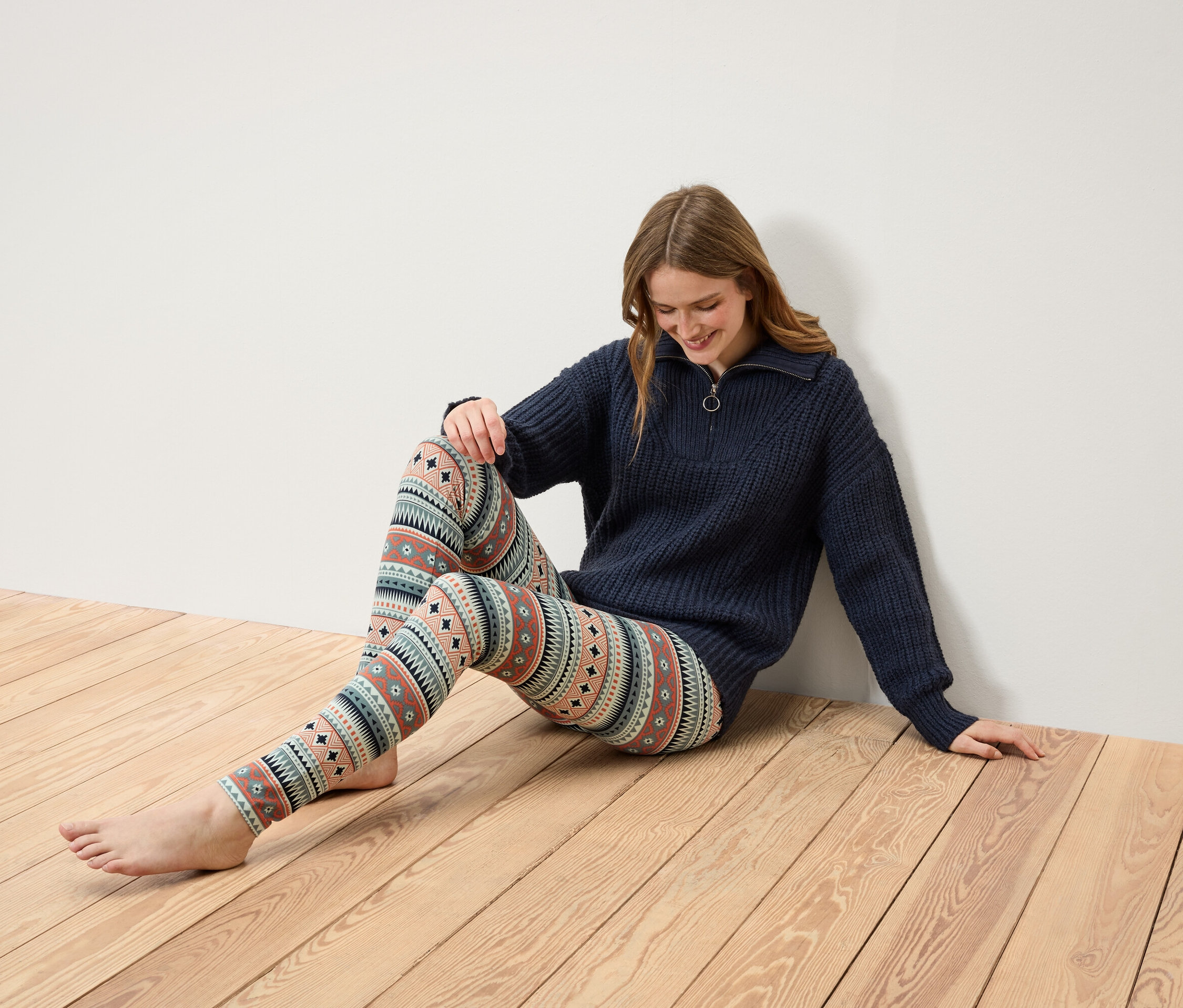Nő ül egy fa padlón, sötétkék, cipzáras pulóvert és mintás leggingst visel.