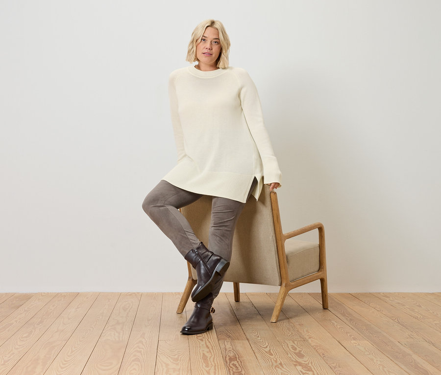 Egy szőke nő pózol egy krémszínű kötött pulóverben, és egy széknek támaszkodik.