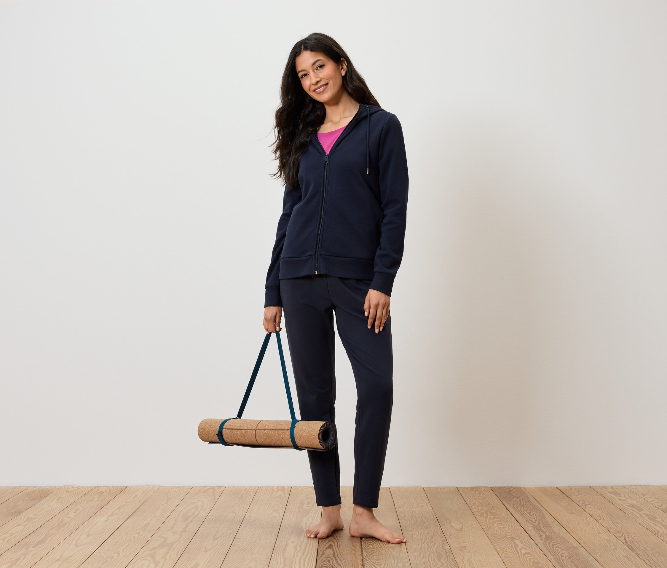 Nő jógamatracot tart, sötétkék tréningruhát és rózsaszín trikót visel.