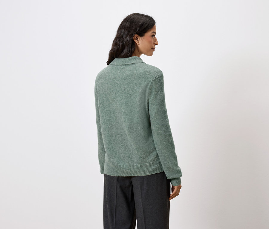 Hátulról látható egy sötét hajú nő, aki zöld pulóvert és sötét nadrágot visel.