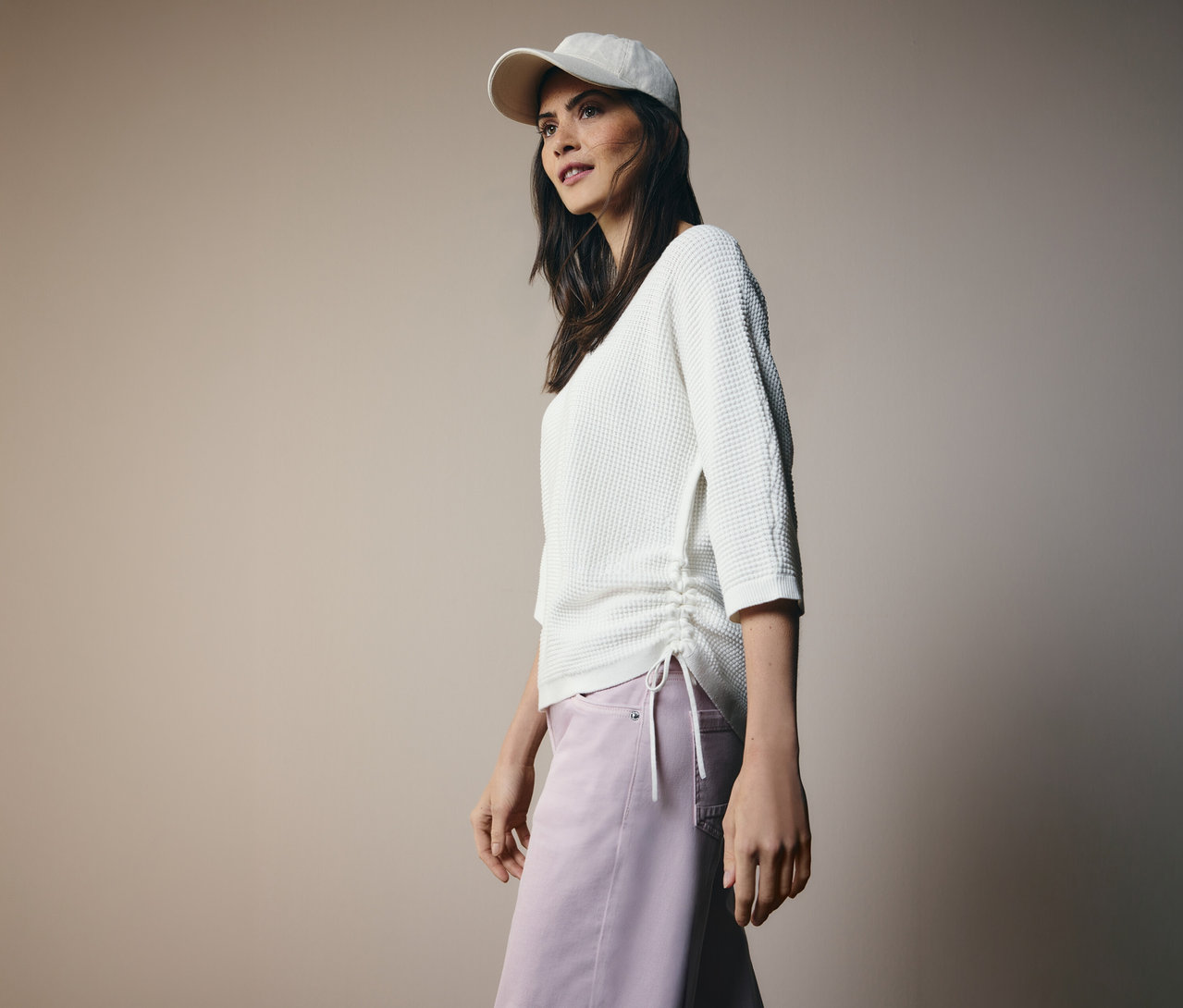 Nő baseballsapkában, oldalt húzózsinóros CECIL finomkötésű pulóverben és rózsaszín nadrágban.